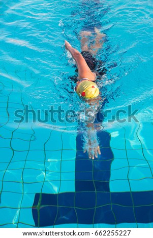 A woman in black swim suits and yellow cap is swimming in freestyle stroke (front crawl) in a swimming pool with clear and blue water.