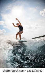 Woman In Black Swimming Vest Balancing On The Wake Board On High Wave Of Motorboat Against Blue Sky