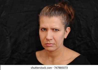 Woman In Black T-shirt On Black Background. Her Face Is Angry And Upset. Her Hair Starts To Be Gray