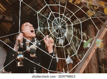 Woman In Black Witch Halloween Masquerade Costume With Skull Candle Holder In Hand Making Spooky Face And Pretending To Be Stuck In Spiderweb While Standing Inside Old Wooden House On Cold Autumn Day