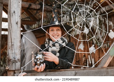 Woman In Black Witch Halloween Masquerade Costume With Skull Candle Holder In Hand Making Spooky Face And Pretending To Be Stuck In Spiderweb While Standing Inside Old Wooden House On Cold Autumn Day