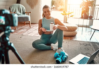 Woman blogger in sportswear sitting on the floor with dumbbells and a laptop and showing a jar of sports nutrition proteins to the camera at home in the living room. Sport and recreation concept. - Powered by Shutterstock