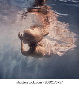     A Woman With Blond Hair In A White Dress Swims Underwater As If Flying In Zero Gravity                           