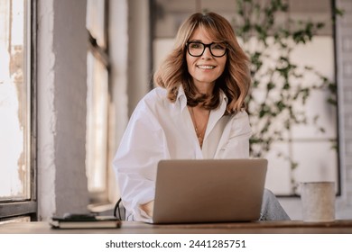 A woman with blonde hair is sitting at home in the kitchen and using a laptop. Mid aged businesswoman working from home and looking at camera and smiling.  - Powered by Shutterstock