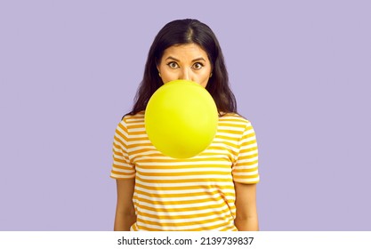 Woman blowing up balloon. Pretty long haired brunette girl with increased lung capacity standing isolated on lilac background, inflating yellow balloon and looking at camera with funny face expression - Shutterstock ID 2139739837