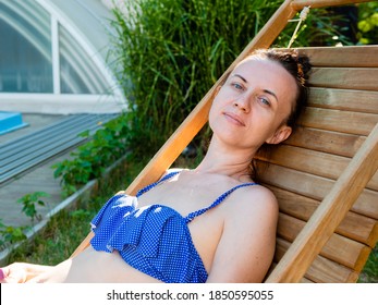 The Woman In The Blue Bathing Suit Rests On A Wooden Lounger In The Backyard And Enjoy The Beautiful Weather.