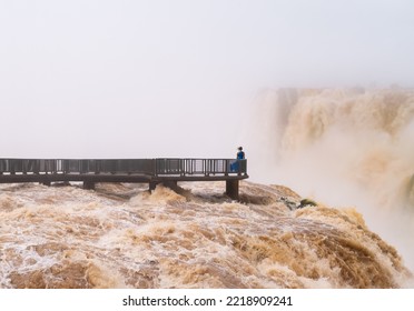 Woman In Blue Dress Enjoys Iguazu Falls From The Viewpoint On The Walkway Between Brazil And Argentina In South America - Adventure And Travel Concept.

