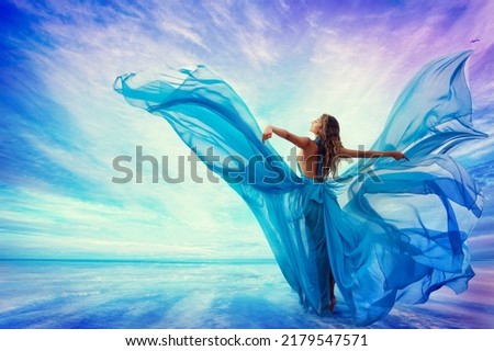 Woman in Blue Dress Flying on Wind looking at Sky and Sea. Beautiful Model Arms outstretched enjoying Freedom at Beach Summer Resort Rear view. Artistic Women Silhouette in Fantasy Gown as Butterfly