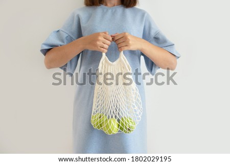 a woman in a blue dress holds a string bag with green apples