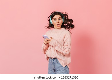 Woman With Blue Eyes In Surprise Looks At Camera On Pink Background. Girl In Knitted Sweater Listens To Music In Headphones And Holds IPhone