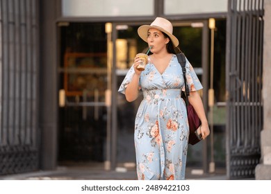 Woman in a blue floral dress and sunhat drinking iced coffee while walking outside a building. She carries a backpack and has a relaxed, casual demeanor.  - Powered by Shutterstock