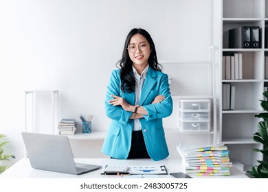 A woman in a blue jacket stands in front of a desk with a laptop and stacks of papers - Powered by Shutterstock