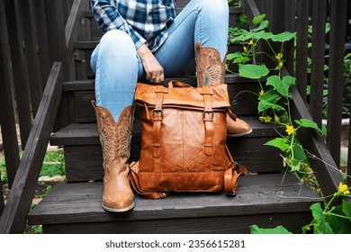 woman in blue jeans and cowboy boots sits on the steps and holds a leather backpack. Close-up.