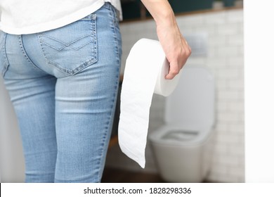 Woman In Blue Jeans Stands With Back And Holds Roll Of Toilet Paper In Her Hand In Front Of Toilet Close-up. Frequent Urination Urology Concept.