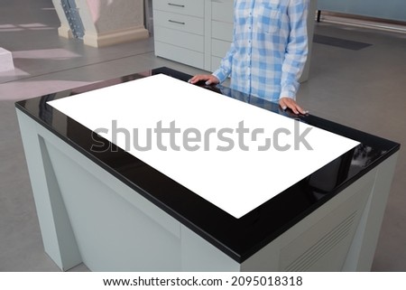 Woman in blue plaid shirt looking at blank interactive touchscreen white display of electronic table kiosk at exhibition or museum. White screen, mock up, copyspace, template, technology concept.