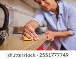 A woman in a blue shirt is cleaning her kitchen counter using a yellow cloth. She seems focused on maintaining a tidy and hygienic home environment. The scene reflects everyday chores.