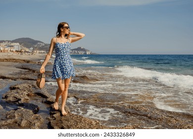 A woman in a blue sundress savors a warm seaside walk, her sandals in hand and the sea stretching out before her - Powered by Shutterstock