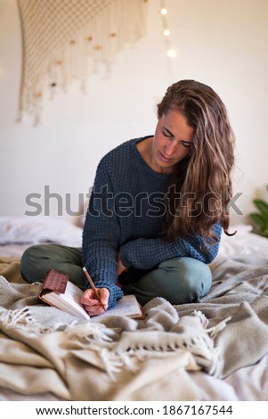 Woman in blue sweater journaling in leatherbound notebook whilst sat on bed
