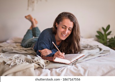 Woman In Blue Sweater Lying On Her Bed Writing In Her Leatherbound Journal