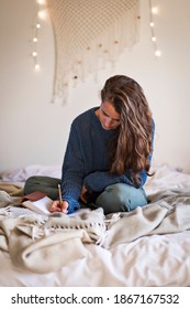 Woman In Blue Sweater Sat On Bed Writing In Her Leatherbound Journal With A Pencil