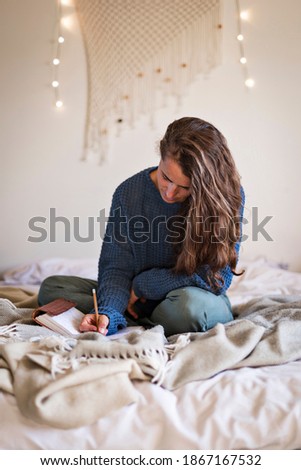 Woman in blue sweater sat on bed writing in her leatherbound journal with a pencil