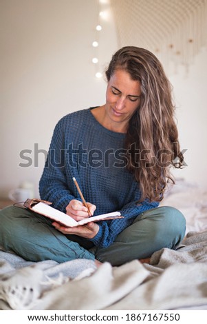 Woman in blue sweater writing in leatherbound journal whilst sat on her bed