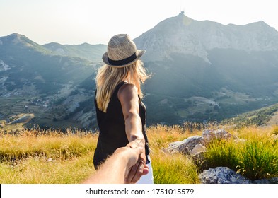 Woman in blue t-shirt and hat holding man by hand in the mountains at glowing sunset. - Powered by Shutterstock