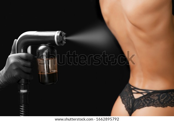 Woman body paint\
instant tan spray skintone with airbrush in professional beauty\
salon on black\
background.