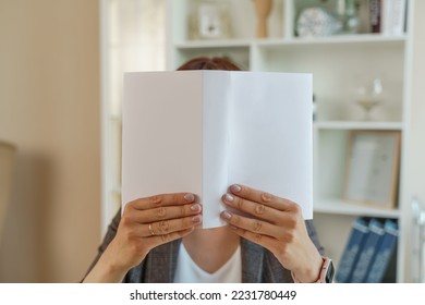 Woman book. Portrait of a girl with an open book in front of her face, she holds it with both hands against the background of the interior. - Shutterstock ID 2231780449