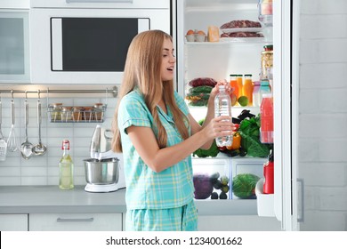 Woman with bottle of water standing near refrigerator in kitchen