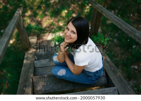 A woman with a bouquet of white wildflowers is sitting on an old wooden bridge looking up. 