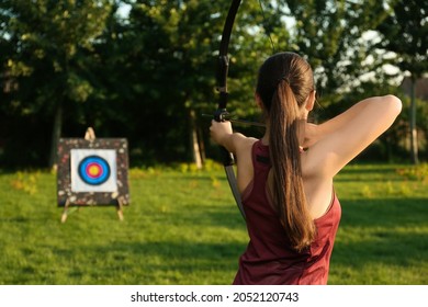 Woman With Bow And Arrow Aiming At Archery Target In Park, Back View