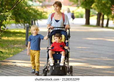 Woman with a boy and a disabled girl in wheelchair walking in park at summer. On baby legs orthosis. Child cerebral palsy. Accessible urban environment for disabled person. Kids with special needs.