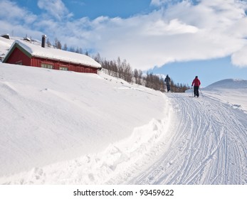 A woman and a boy skiing in a groomed ski track up hill passing a hunting lodge at easter in the norwegian mountains