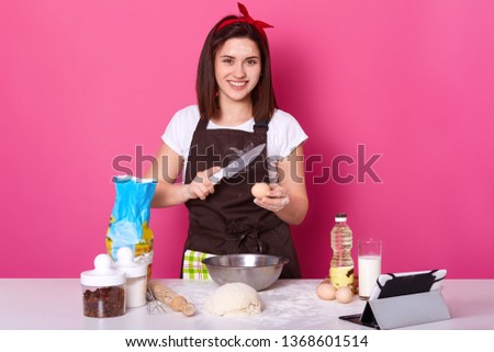 Woman brakes egg with knife, puts it into plate of flour, needs some more dough for preparing hot cross buns, ready for kneading dough, looks happily at camera, posing isolated over pink studio wall.