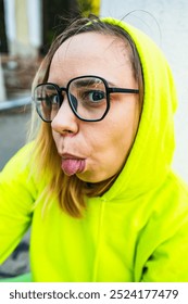 A woman in a bright yellow hoodie playfully sticks out her tongue while posing outdoors on a sunny day