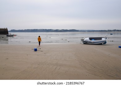 Woman in bright yellow jacket walking on a sandy beach towards the sea at low tide, with small boats moored in the harbor