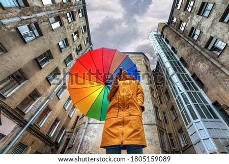 A woman in a bright yellow raincoat with a rainbow umbrella standing in an old courtyard well and looking at the stormy sky. Saint Petersburg, Russia