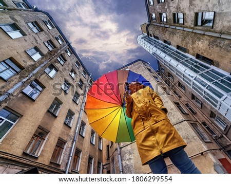 A woman in a bright yellow raincoat with a rainbow umbrella standing in an old courtyard well and looking at the dark stormy sky. Saint Petersburg, Russia