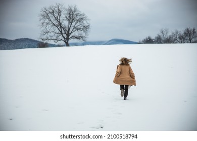 Woman In Brown Coat Runs On A Snow Field. Mountains In The Distance. Free Spirit Girl. Winter Season. High Quality Photo