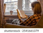 Woman with brown hair is sitting in soft armchair, wrapped in plaid, and reading book. Outside window are winter trees in snow. Cozy solitude. Back view.