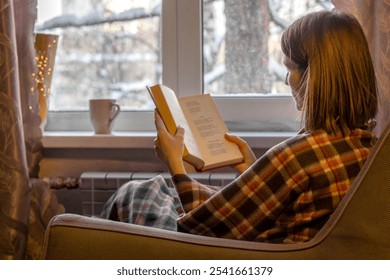 Woman with brown hair is sitting in soft armchair, wrapped in plaid, and reading book. Outside window are winter trees in snow. Cozy solitude. Back view. - Powered by Shutterstock