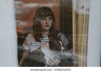 Woman with brown hair and a striped shirt sits by a rainy window, holding a Boston Terrier. Rainy day, woman and dog, peaceful moody and thoughtful moment indoors. Rainy day moody photo. - Powered by Shutterstock