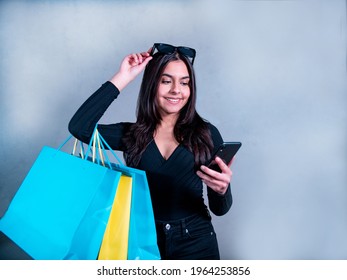 Woman with brown hair, sunglasses, in black jeans and jersey carries blue and yellow shopping bags while she looks at her phone. 