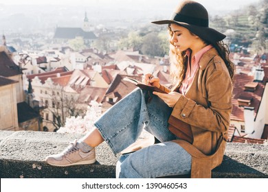 Woman in brown hat writing in the diary in view point with amazing view. Cute woman taking notes and wirting in notebook.
