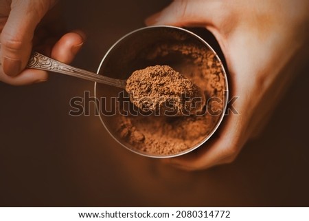 A woman with a brown manicure on her nails holds a jar of fragrant crumbly cocoa powder in her hands and scoops a portion with a silver teaspoon. A delicious drink in the morning. The cooking process.