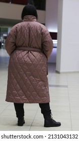 Woman In Brown Winter Waterproof Coat With Black Pants Holding Hands In Pockets Rear View.