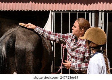 Woman brushing a dark horse with child girl observing near a stable. Routine on a farm, taking care of animals in the countryside - Powered by Shutterstock