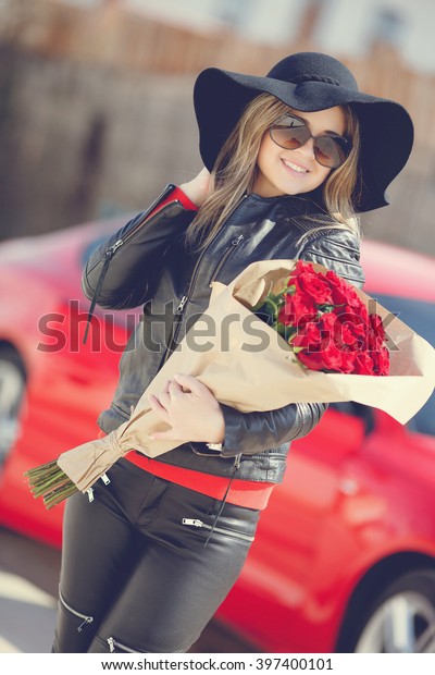 woman with bunch of red flowers near red car full of\
gift boxes. roses, woman and car. car trunk full of presents. happy\
woman. holiday. Sexy woman with bouquet. Happy woman near luxury\
red car