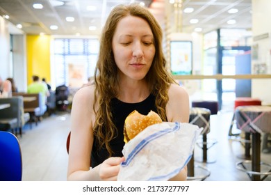 Woman With Burger In A Fast Food Restaurant. Woman Opening Wrapping Paper, Holding, Biting The Burger And Savouring Food. Fast Snack On The Go